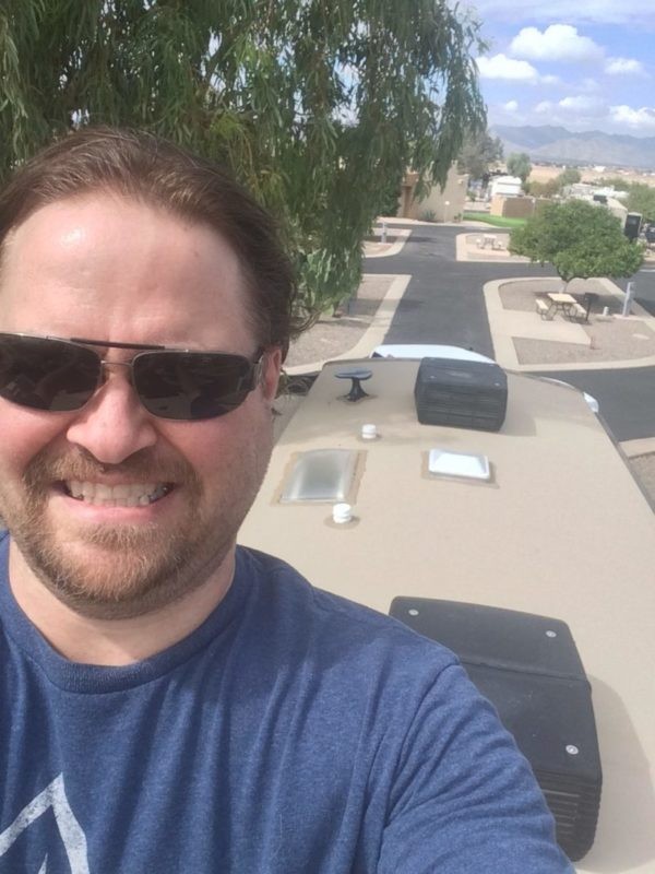A white man with brown hair and a short beard wearing sunglasses and a blue shirt is looking at the camera, with an RV roof behind him and the view of an RV park in the distance.