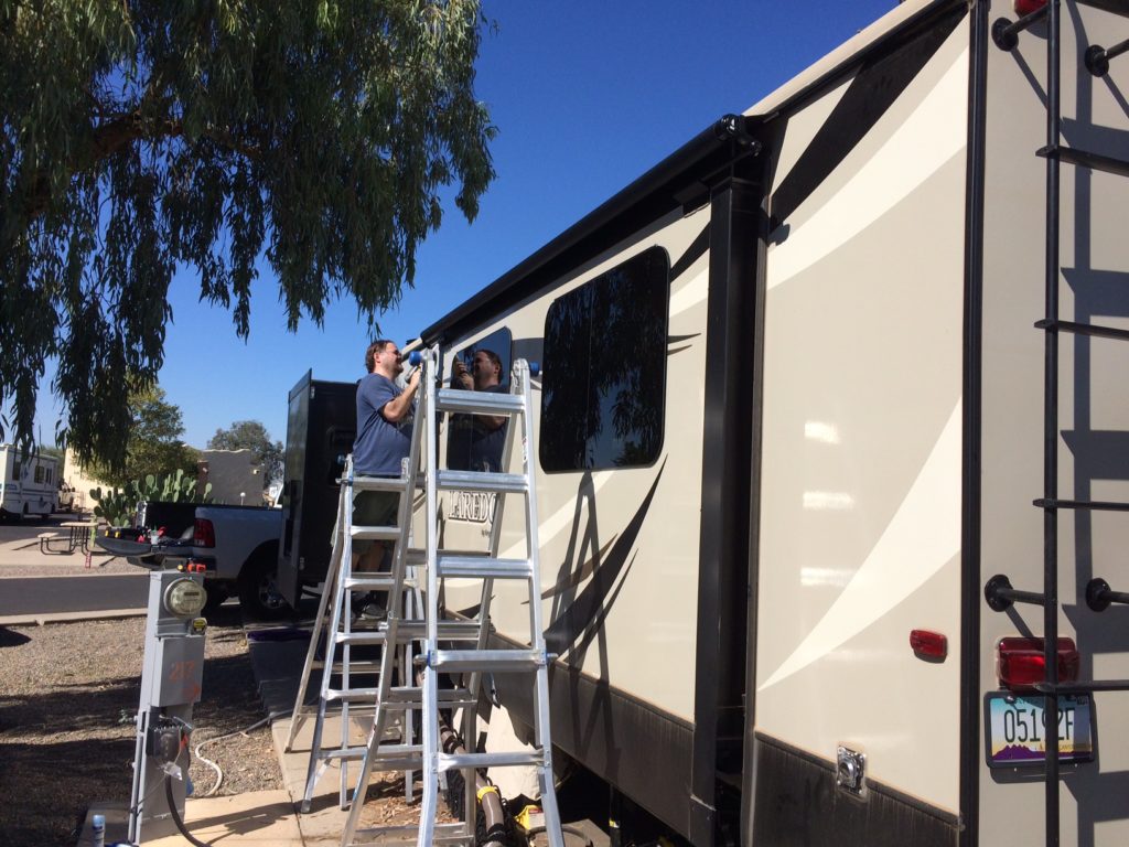 The side of an RV, with two ladders next to a slide-out. A man is standing on the ladder farthest from the camera, working on the slide.