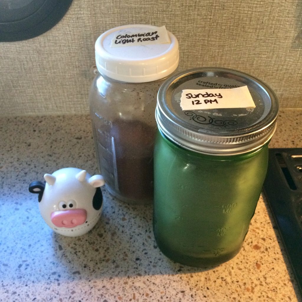 Two mason jars on a counter next to a kitchen timer shaped like a cow. One of the jars is green glass with a sticker on the lid labeling it Sunday at 12PM, and the other jar is half-full of ground coffee with a label describing the contents.
