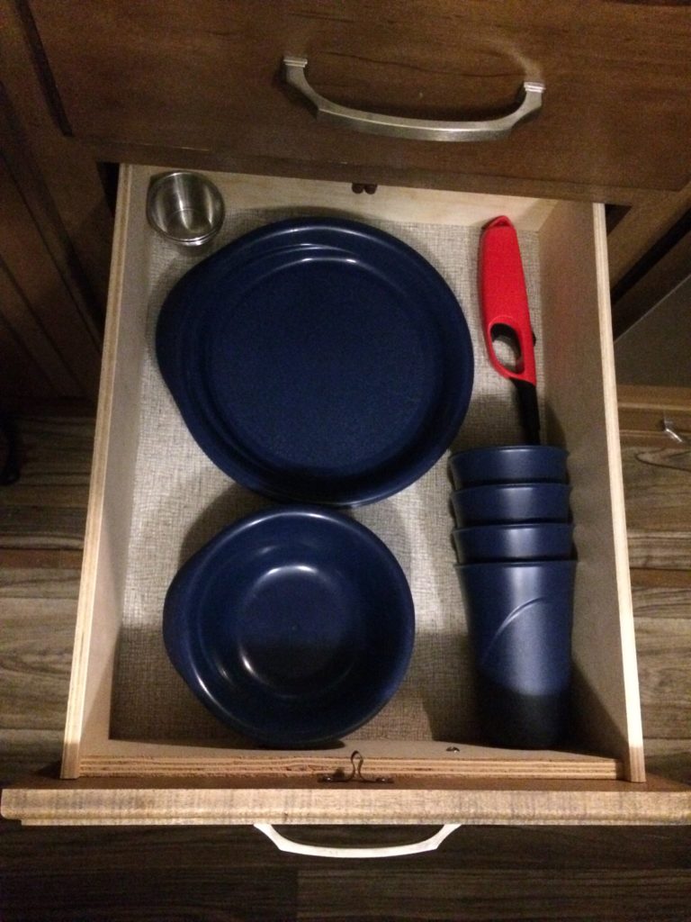 An open drawer, with a stack of plates, bowls, and cups inside, as well as a long-handled lighter, and a small stack of stainless steele ramekins.