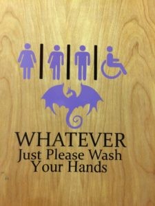 Bathroom door with a male figure, female figure, the half-skirt figure intended to represent trans people, a wheelchair, and a dragon. The words "Whatever just please wash your hands" are underneath the images.