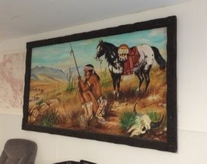 A sad native american man in traditional garb and carrying a spear, next to a horse in traditional garb. He is watching a caravan of covered wagons in the distance, looking mournful.