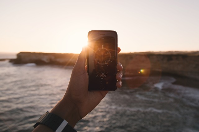 A hand holding a smart phone with a compass app pointed towards the horizon with the sun setting in the distance
