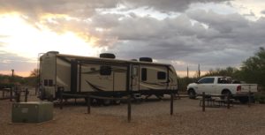 A white pickup truck parked in front of an RV trailer, with the sun setting and clouds in the distance.