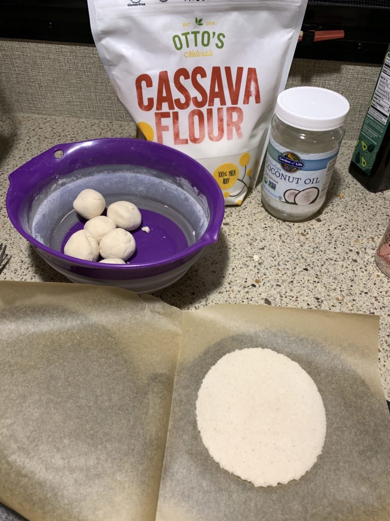 a bag of cassava flour, a jar of coconut oil, a bowl of small balls of dough, and a tortilla on parchment paper on a tortilla press.