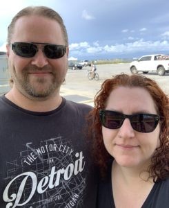 a man and a woman wearing sunglasses and smiling at the camera. a beach is behind them.