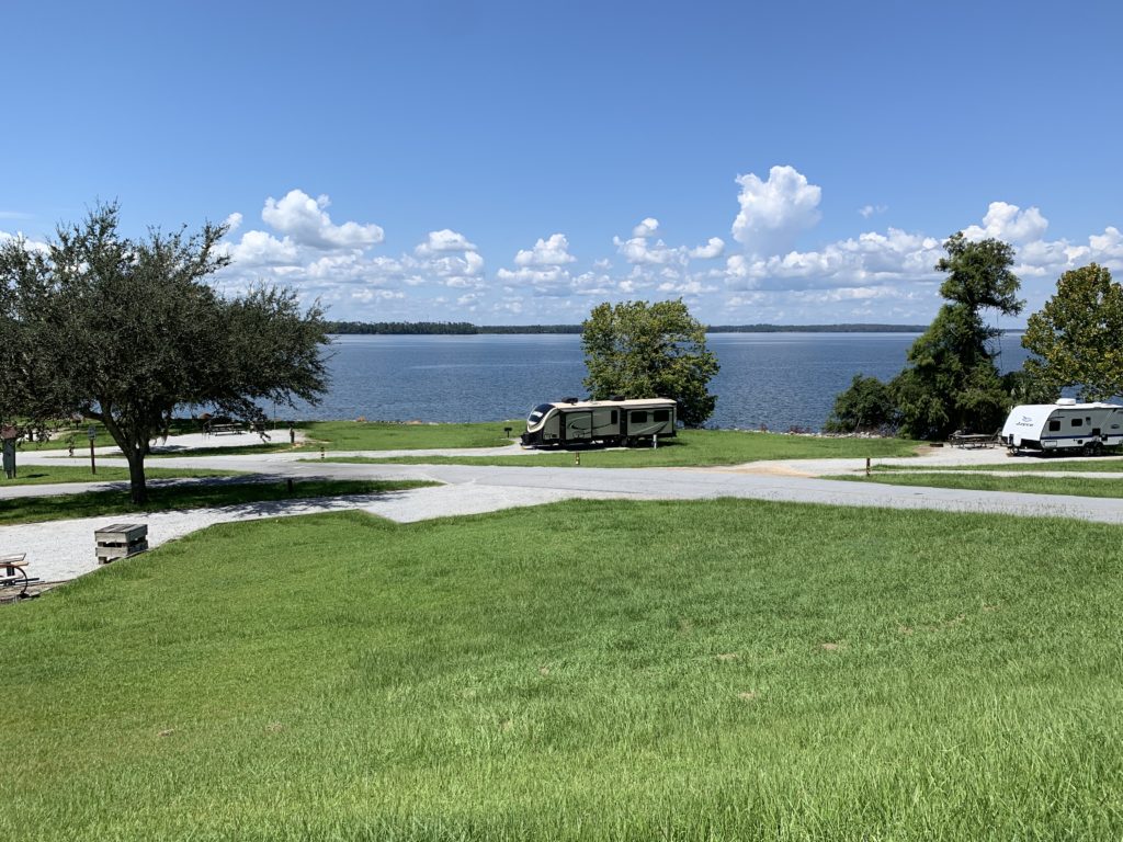 a view from on top of a hill, looking down at an RV parked along a large, calm lake, with lots of grass, a few trees, and very blue skies.