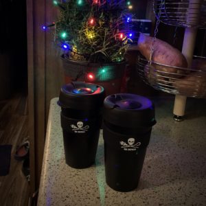 two black reusable coffee cups in front of a tiny lit up tree on a counter.