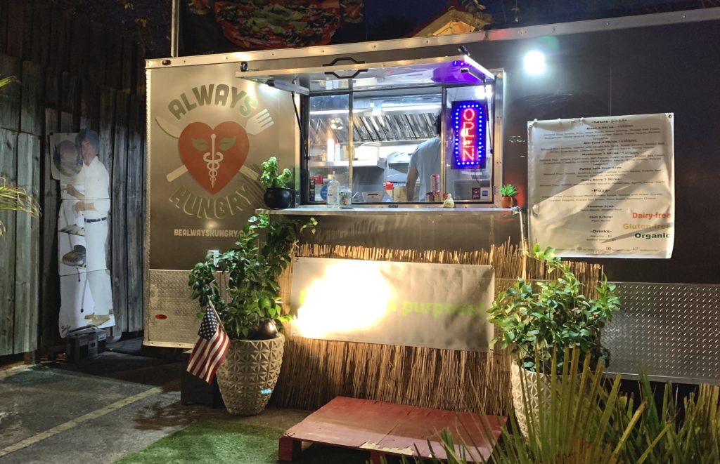 a food truck/trailer with two plants out front, a pallet in front of the window as a step, and a cardboard cutout of Bob Ross in the background.