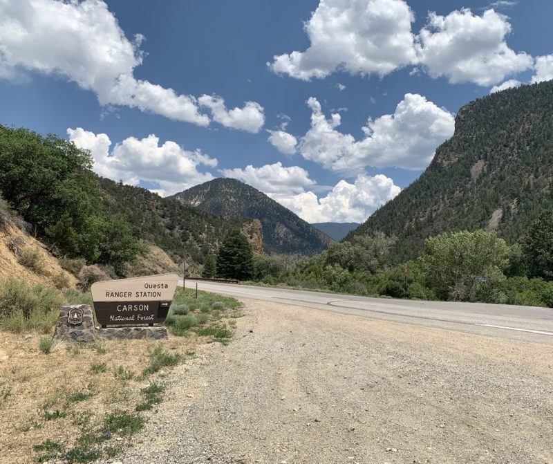 Photo of the sign for the Questa Ranger Station with mountains in the distance, blue skies, and big puffy clouds.