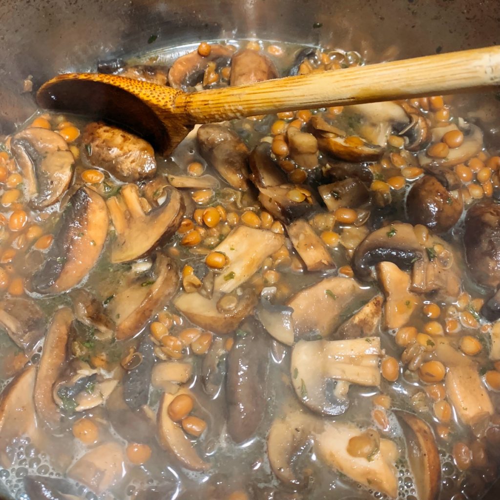 A pot of steaming lentils and mushrooms with a wooden spoon resting on the edge of the pot 
