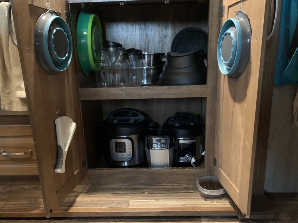 An under-counter cabinet with collapsible bowls hanging on each cupboard door, with two instant pots and a blender on the bottom shelf, and an instant pot air fryer, two pots, blender cups and two glass bottles on the top shelf. a third larger collapsible bowl hangs inside the cabinet, and a jar opener hangs on the lower half of one door. a small bowl of cat food sits on the floor under the cabinet doors.