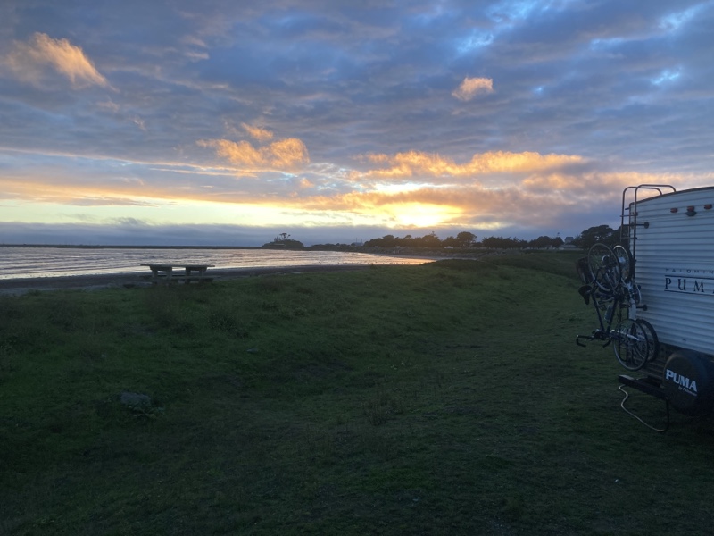 Sun setting behind the clouds at the shore. There is a grassy berm with a picnic table on top, and the crescent city lighthouse can be barely be seen in the distance. 