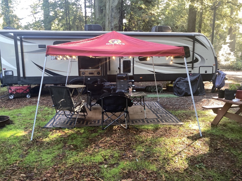 A canopy tent/sun shade over some chairs and an outdoor rug. There is a string of lights hanging from the tent and the tent is red with a picture of the beaver from the Buc-ee’s logo on it. A travel trailer is parked behind the tent. 